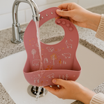 Pink silicone bib with animal prints being rinsed under running water in a sink.