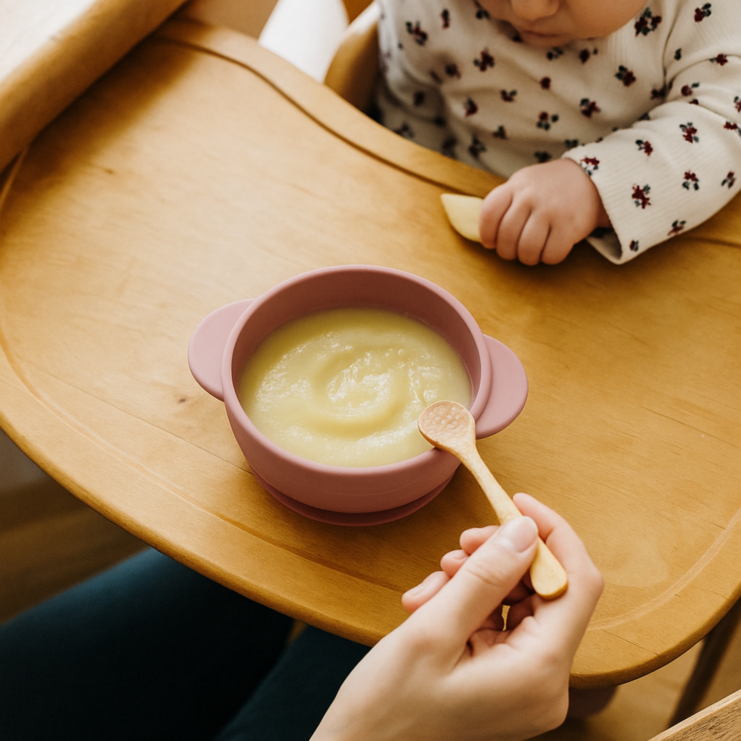 Child in a high chair with a pink bowl of baby food and a wooden spoon, held by an adult's hand.